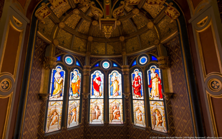 Tall stained-glass window with colorful figures set in a richly painted interior with decorative ceiling.