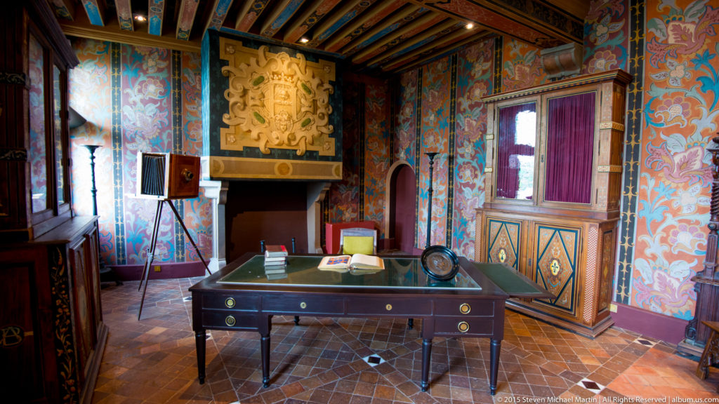 Historic room with desk, patterned walls, painted timber ceiling, carved overdoor, and window alcove.