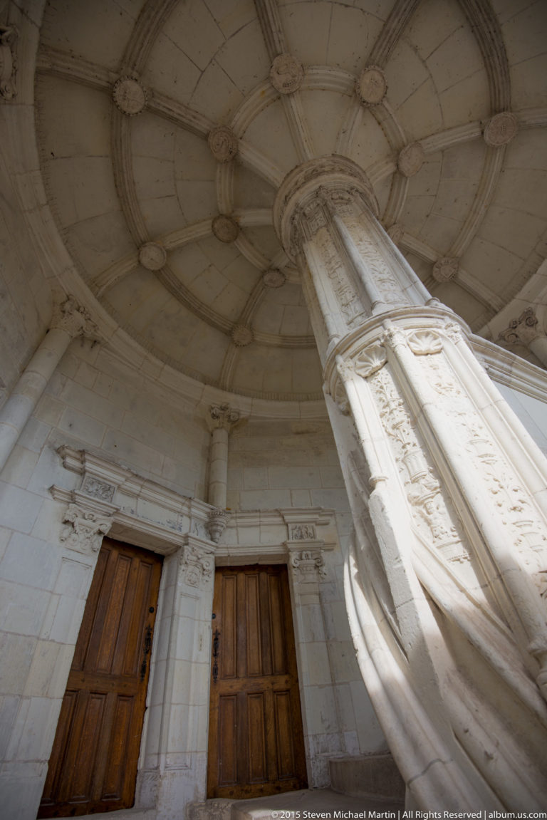 Upward view of a carved stone column rising into a circular ribbed ceiling above wooden doors.