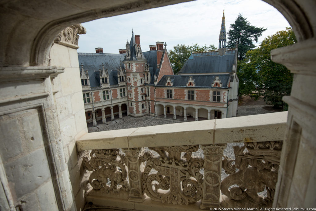 Elevated view across carved stone balustrade toward the brick-and-stone courtyard wing and spire at Blois château.