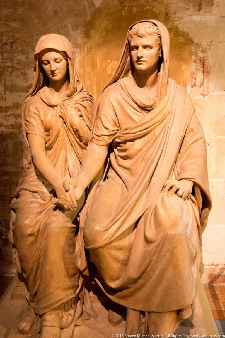 Two seated draped female stone figures, one beside a smaller figure, displayed against a warm-toned wall.