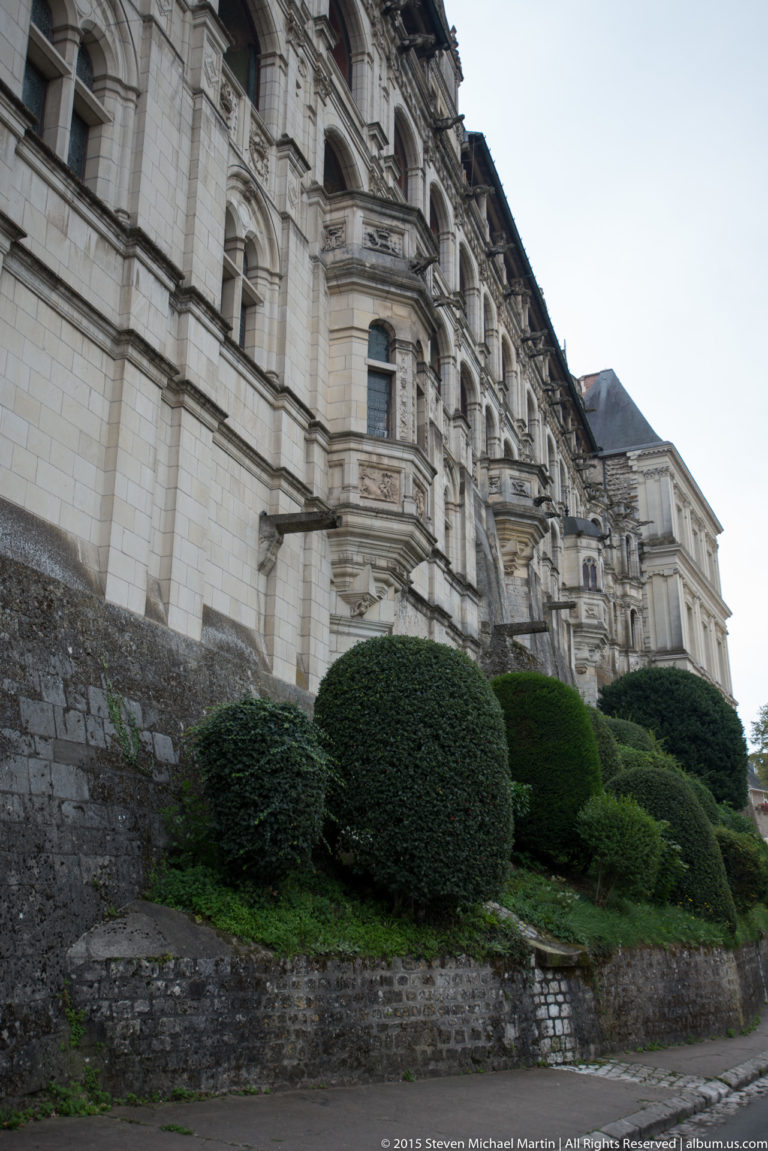 Long pale stone château façade with repeated windows and projecting bays above clipped shrubs on a sloping embankment.