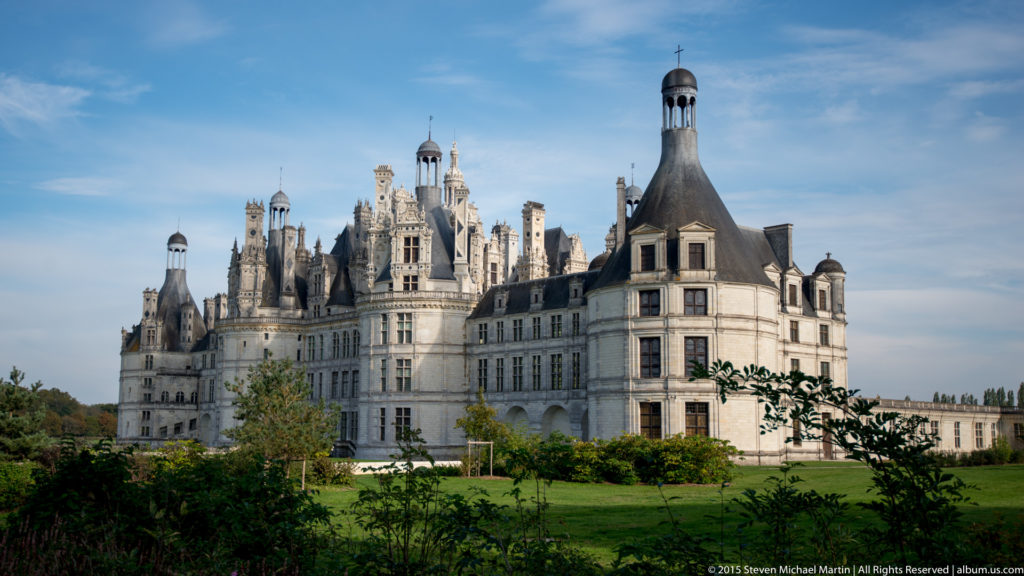Exterior view of Château de Chambord with pale stone façades, corner towers, steep roofs, chimneys, and ornate rooftop lanterns.