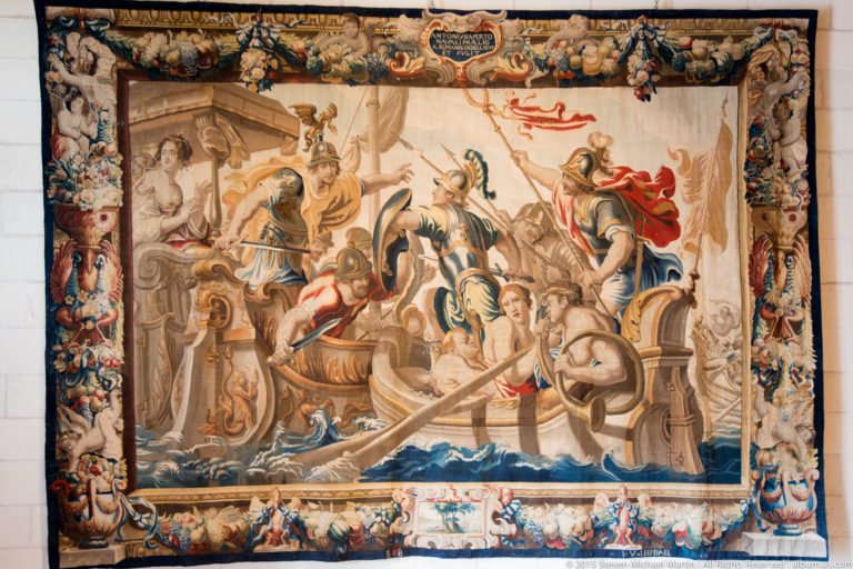 Large historic tapestry with a crowded figurative battle scene, horses, water, and an elaborate decorative border.