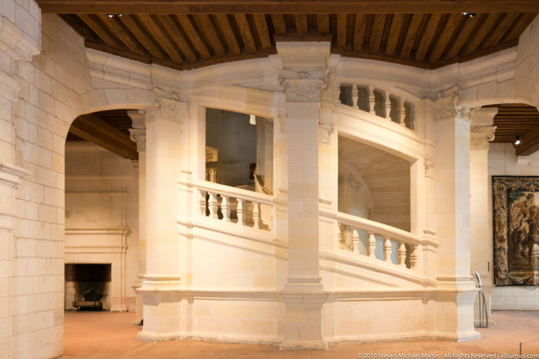 White stone staircase and balustraded gallery beneath timber ceiling beams, with arches and a tapestry in the adjoining room.