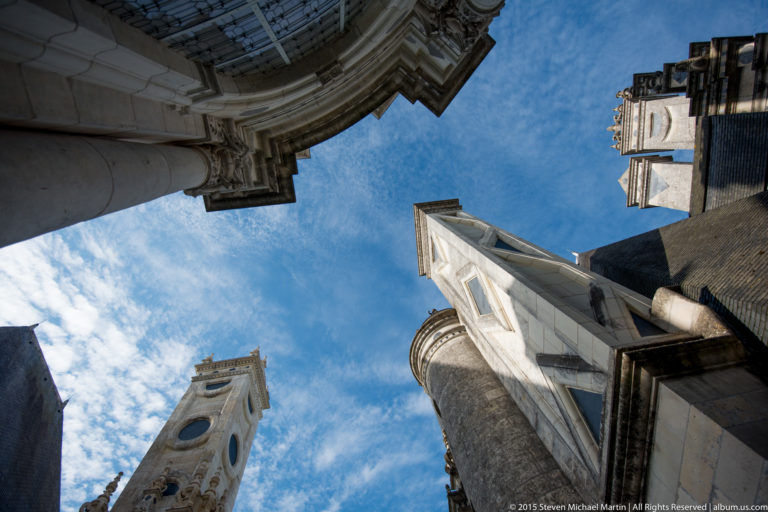 Upward-looking view between Château de Chambord towers, chimneys, slate roofs, and carved stone projections against blue sky.