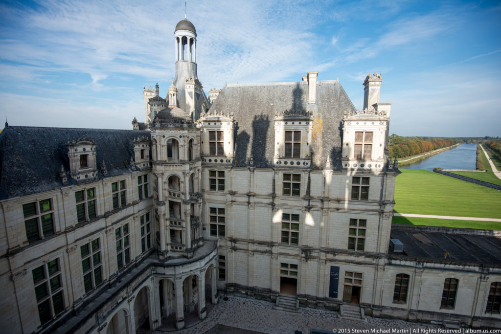 Elevated view over Château de Chambord courtyard roofs toward green lawns and a long straight water feature.