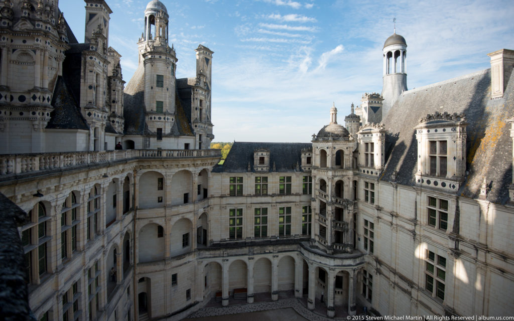High-angle view into Château de Chambord courtyard with stacked arcades, windows, towers, and strong sun and shadow.