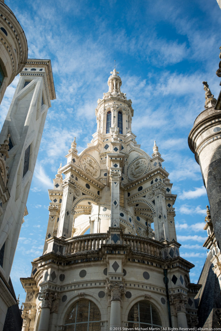 Low-angle close view of an ornate Château de Chambord rooftop tower with columns, circular openings, carvings, and lantern.