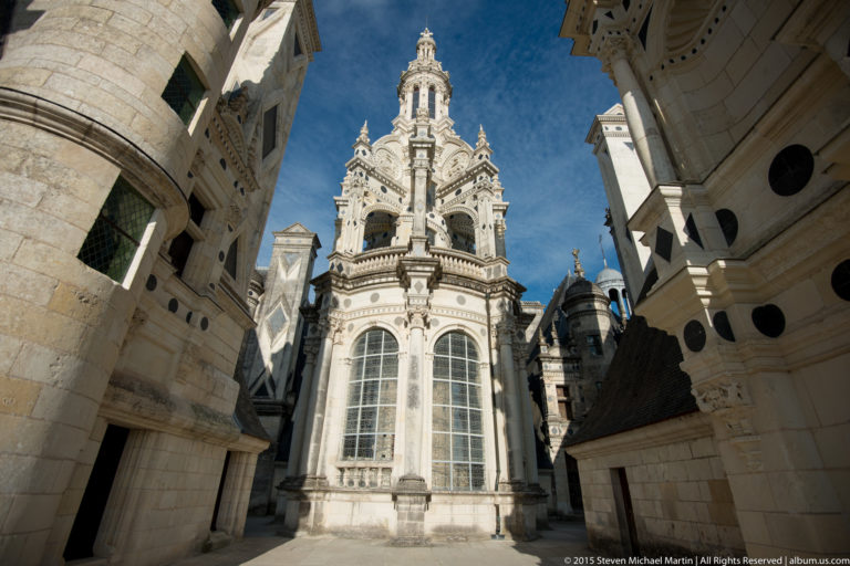 Low-angle view of an ornate tall windowed pavilion with carved stonework and lantern, framed by adjoining Château de Chambord walls.