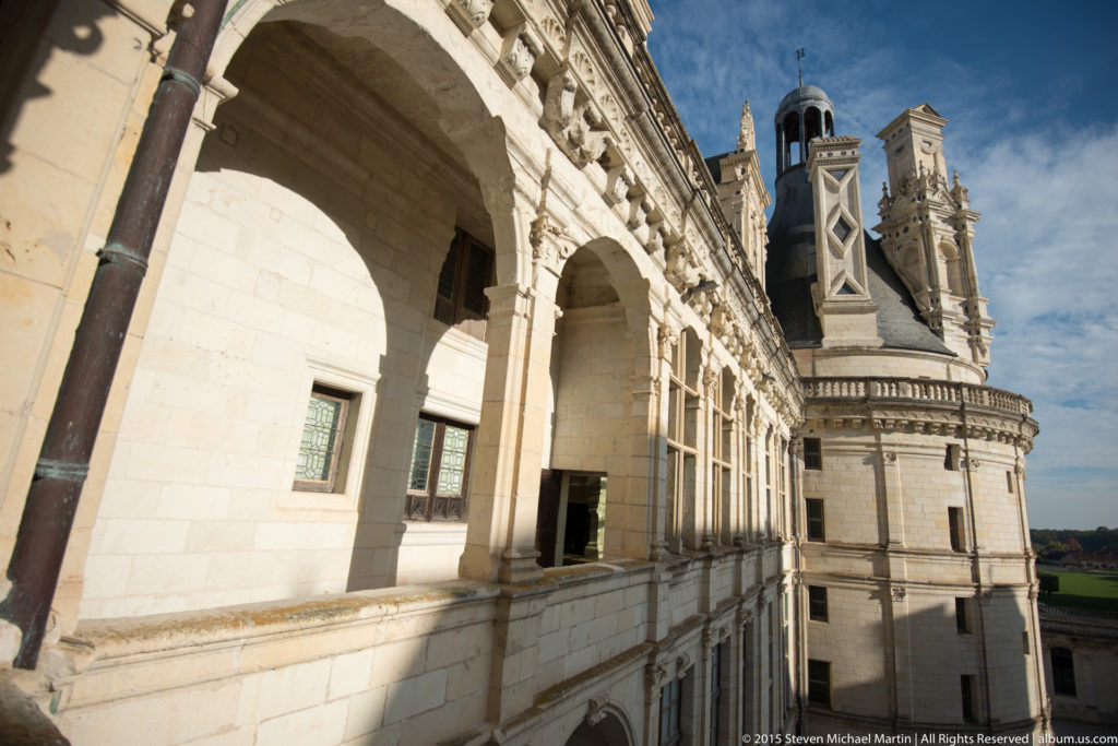 Oblique view along an open stone arcade at Château de Chambord toward a round tower and distant landscape.