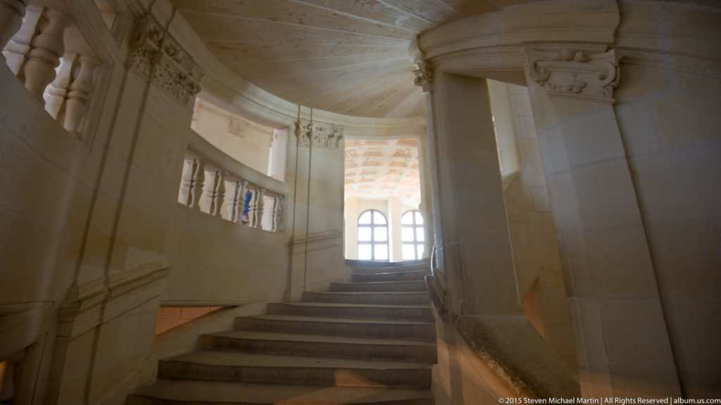Curving stone staircase inside Château de Chambord with thick piers, vaulted ceiling, balustrade, and bright window ahead.