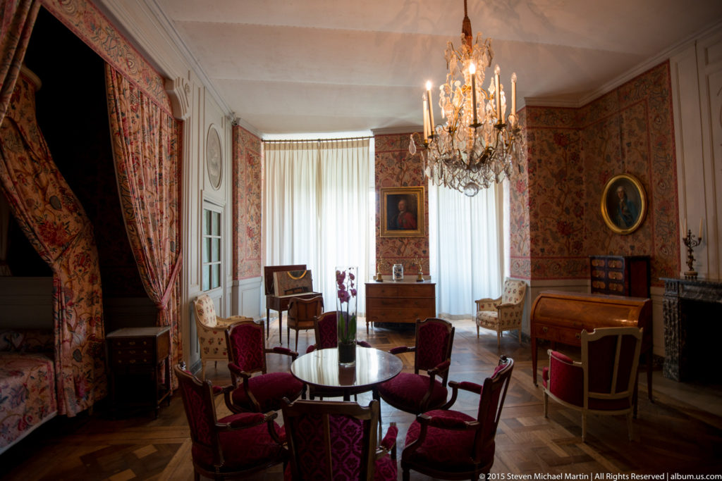 Historic furnished room with patterned red textiles, draped bed or alcove, chairs, tables, portraits, and crystal chandelier.