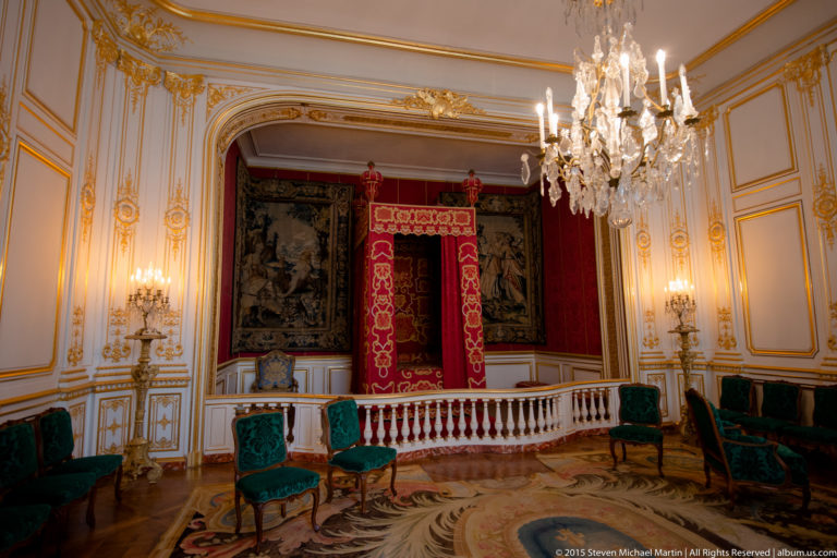 Red canopied state-style bed centered in a gilded room with tapestries, chandeliers, and green upholstered chairs.