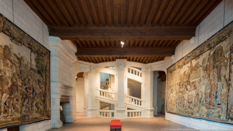 Stone gallery at Château de Chambord with large tapestries on both walls, timber ceiling beams, central staircase, and red bench.