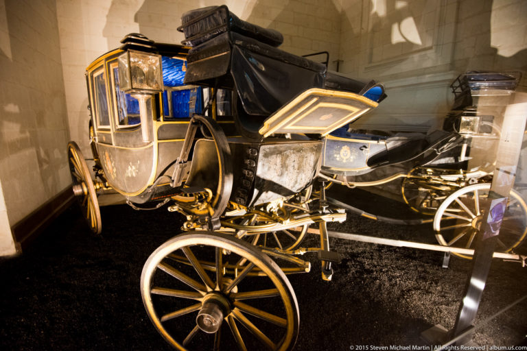 Black and gold historic horse-drawn carriage with glass cabin, large spoked wheels, suspension, and raised driver’s seat.