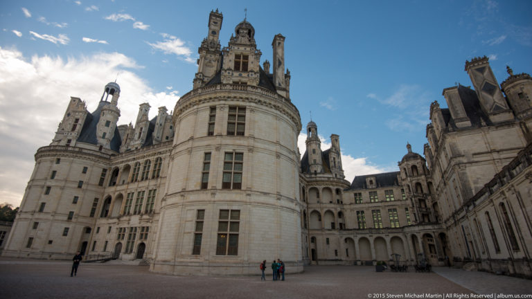 Wide courtyard view of Château de Chambord with rounded tower, tall windows, ornate roofline, and small visitors below.
