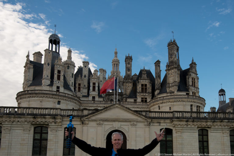 Visitor with arms extended in front of Château de Chambord, with towers, chimneys, and ornate roofline behind.