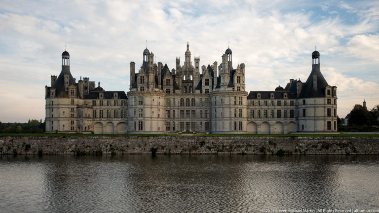 Château de Chambord seen across broad water, with central keep, flanking wings, towers, and soft reflections.