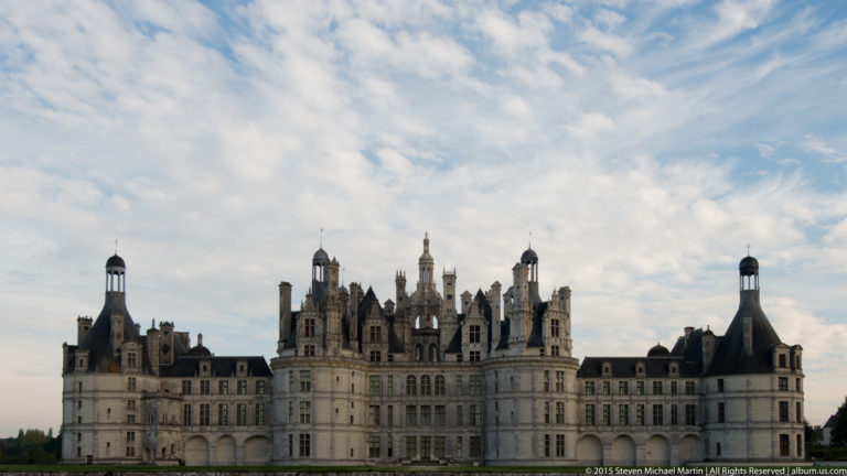 Distant frontal view of Château de Chambord beneath a large cloud-filled sky, with central keep and long wings visible.