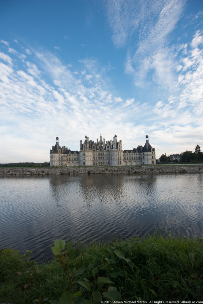 Very distant panoramic view of Château de Chambord across calm water, with low shoreline vegetation and broad blue sky.