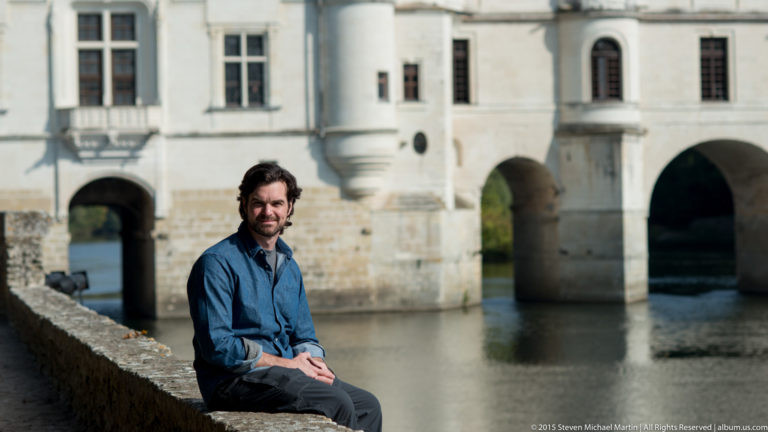 Seated dark-haired man in a blue shirt with the arched Château de Chenonceau gallery blurred behind him.