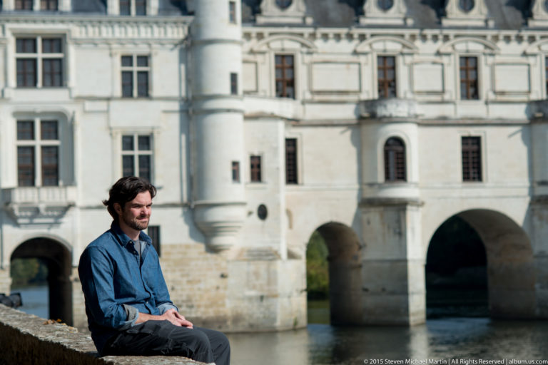 Dark-haired man seated in profile before the softly focused arched façade of Château de Chenonceau.