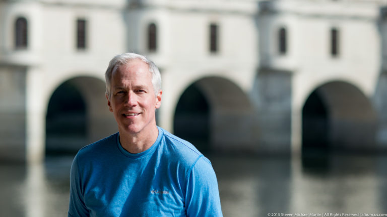 Gray-haired man in a bright blue shirt before blurred river arches at Château de Chenonceau.