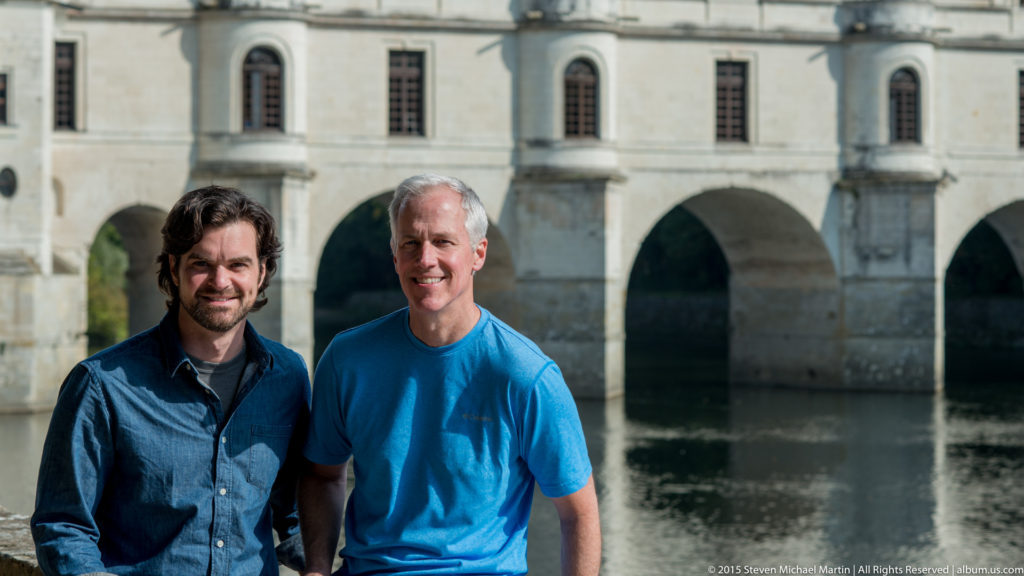 Two men standing together in front of the pale stone gallery and river arches of Château de Chenonceau.