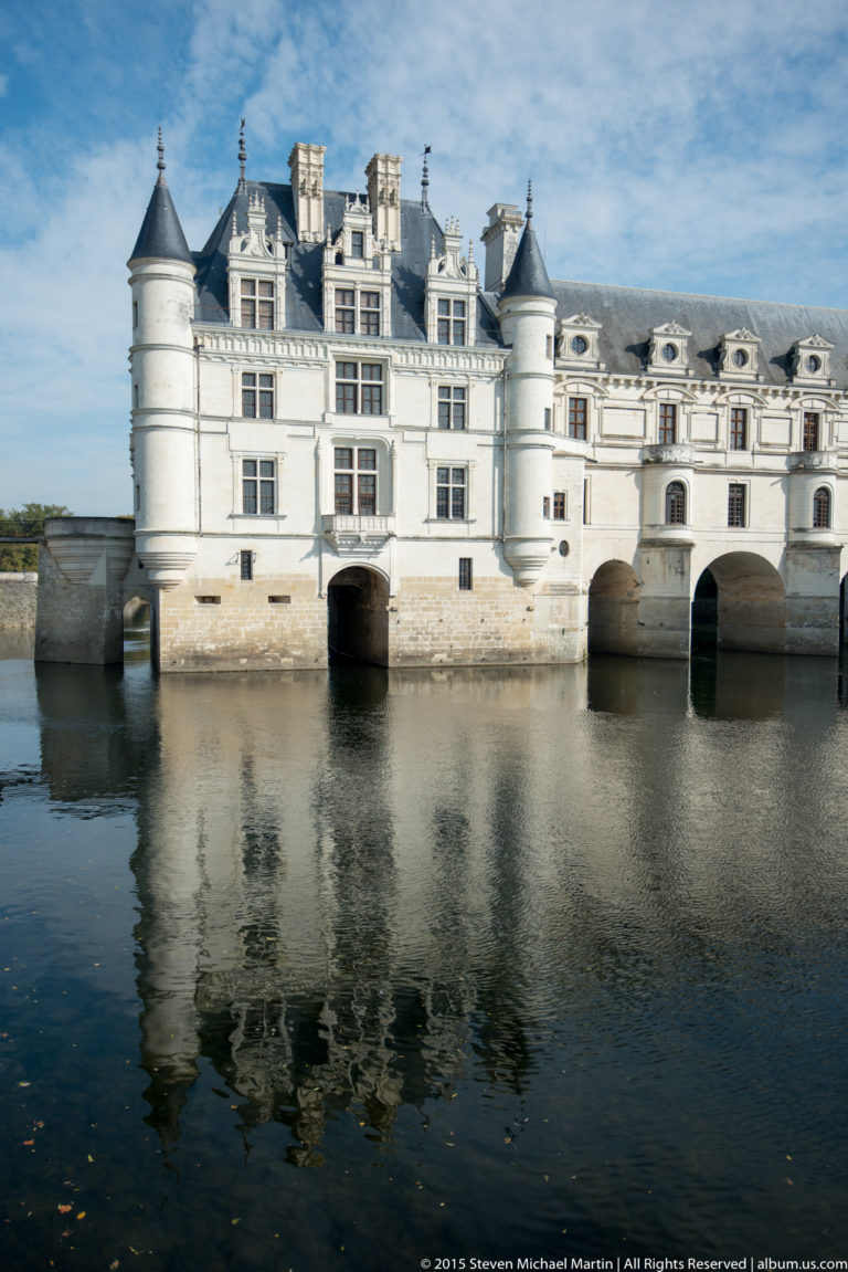 Frontal view of Château de Chenonceau with round turrets, river arches, and a broad reflection in the Cher.