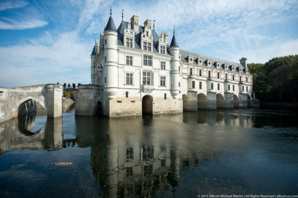Oblique view of Château de Chenonceau beside a low bridge, with turrets and reflections in the River Cher.