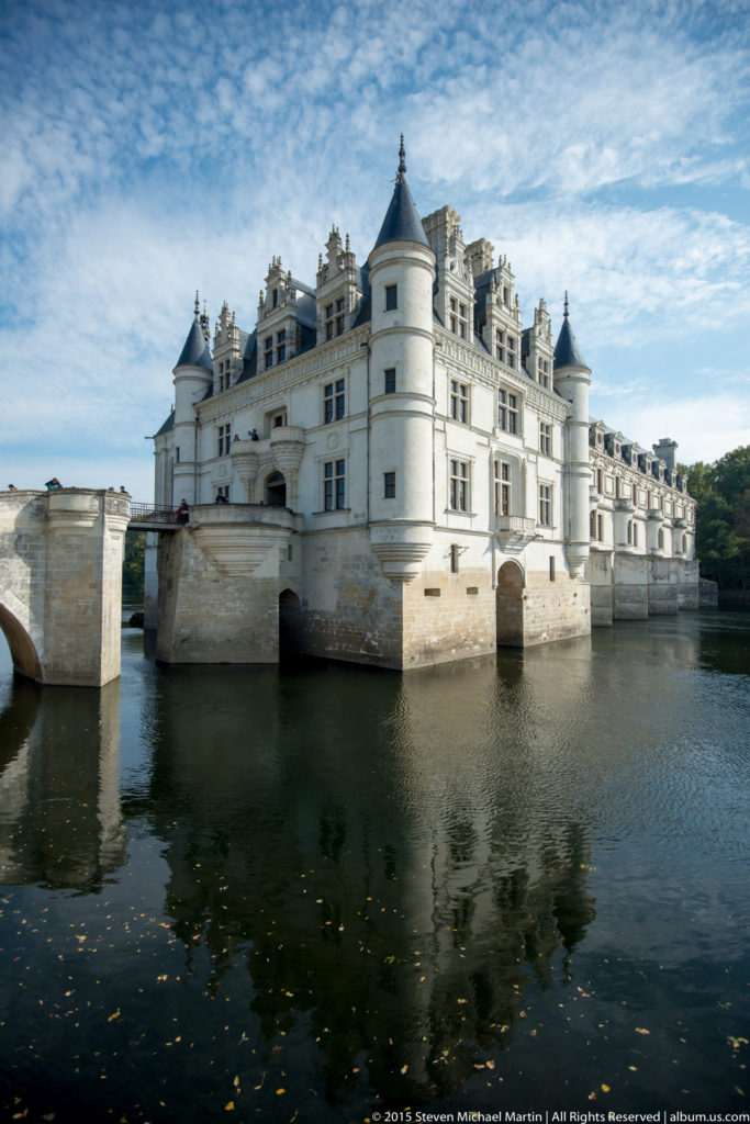 Wide angled view of Château de Chenonceau extending over the River Cher beneath a blue sky.
