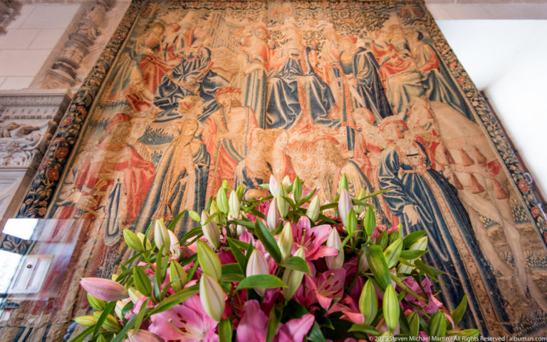 Large historic figurative tapestry behind a foreground arrangement of pink and white lilies inside Château de Chenonceau.