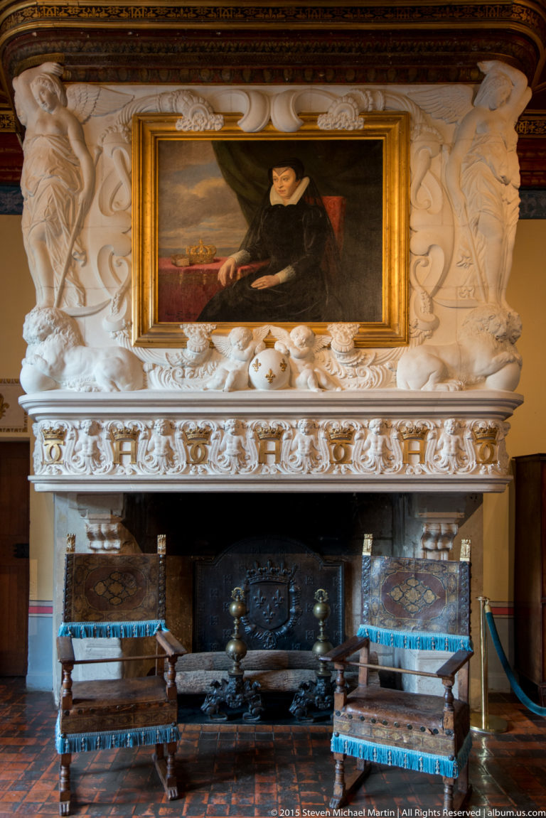 Ornate carved-stone fireplace with framed portrait above and upholstered benches inside Château de Chenonceau.