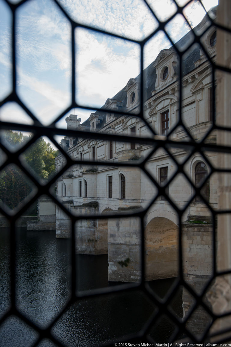 Château de Chenonceau seen through a dark leaded window lattice, with the river and pale façade beyond.