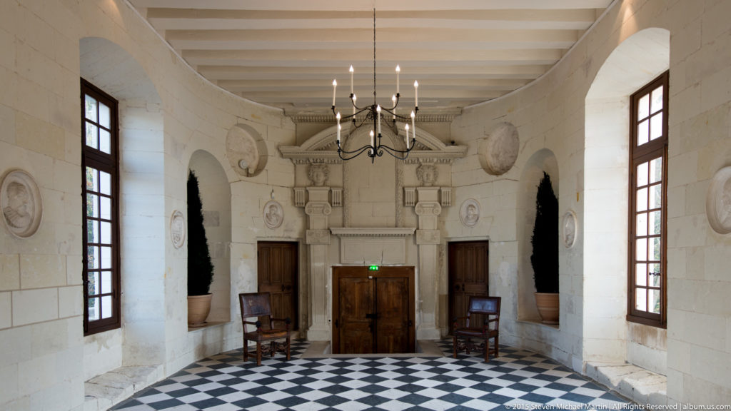 Stone interior hall with black-and-white checkerboard floor, tall windows, wooden doors, and chandelier at Château de Chenonceau.