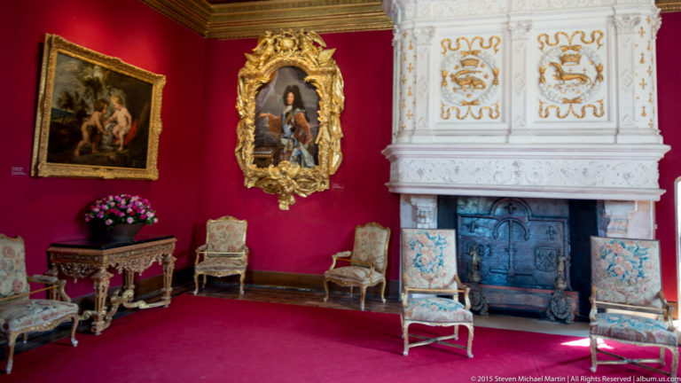 Red-walled salon with gilded paintings, pale carved fireplace, upholstered chairs, and flowers at Château de Chenonceau.