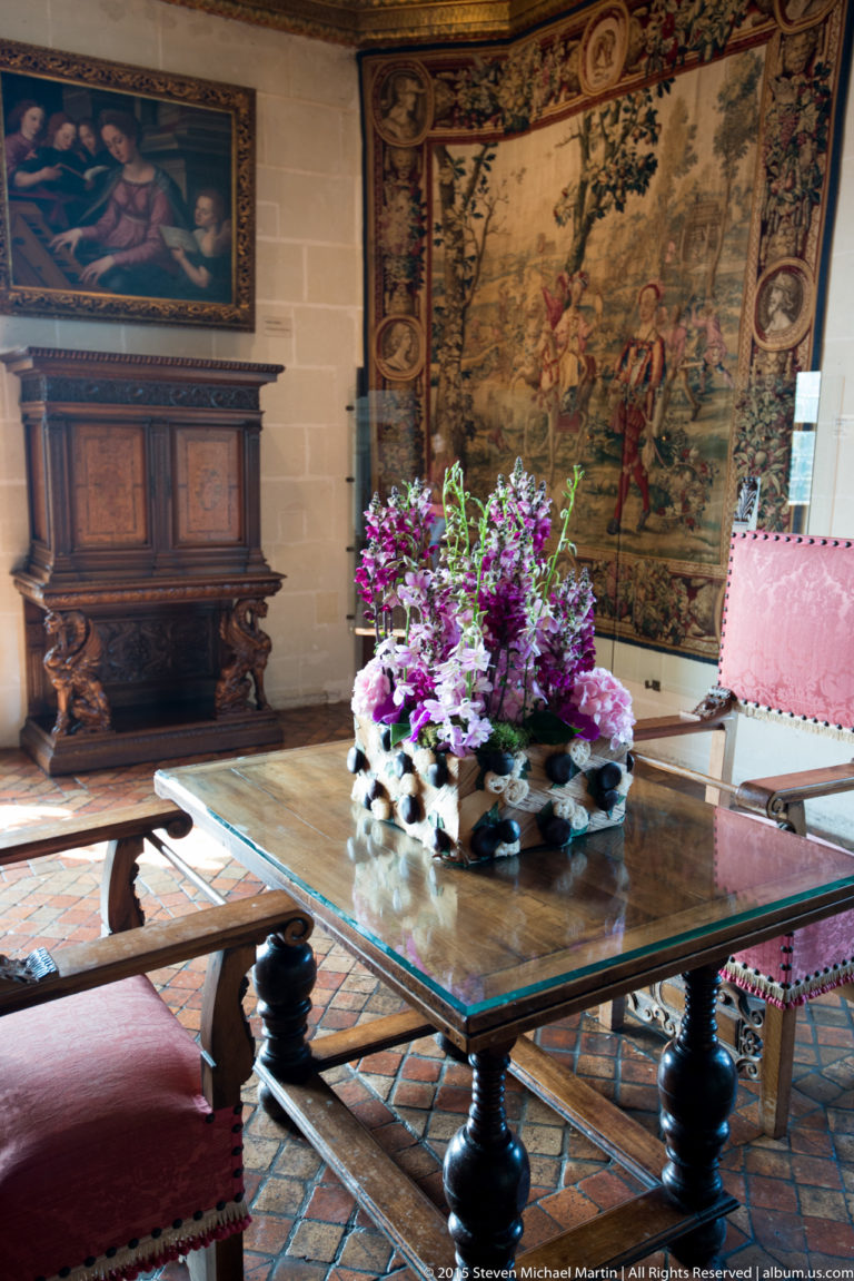 Historic room with large tapestry, carved wooden cabinet, heavy table, upholstered chairs, and purple flowers at Château de Chenonceau.
