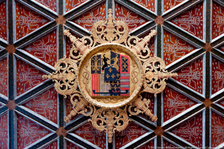 Close view of a gilded heraldic shield and ornate crest set within a red painted coffered ceiling at Château de Chenonceau.