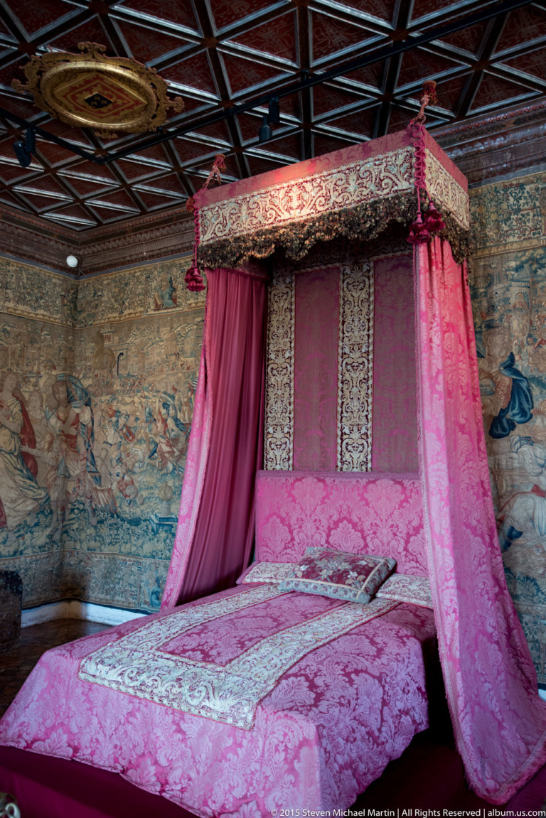 Pink canopied bed with patterned hangings beneath a dark coffered ceiling and tapestry-covered walls at Château de Chenonceau.