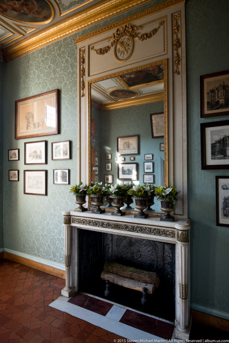 Green-walled room with pale marble fireplace, large mirror, framed pictures, and greenery at Château de Chenonceau.