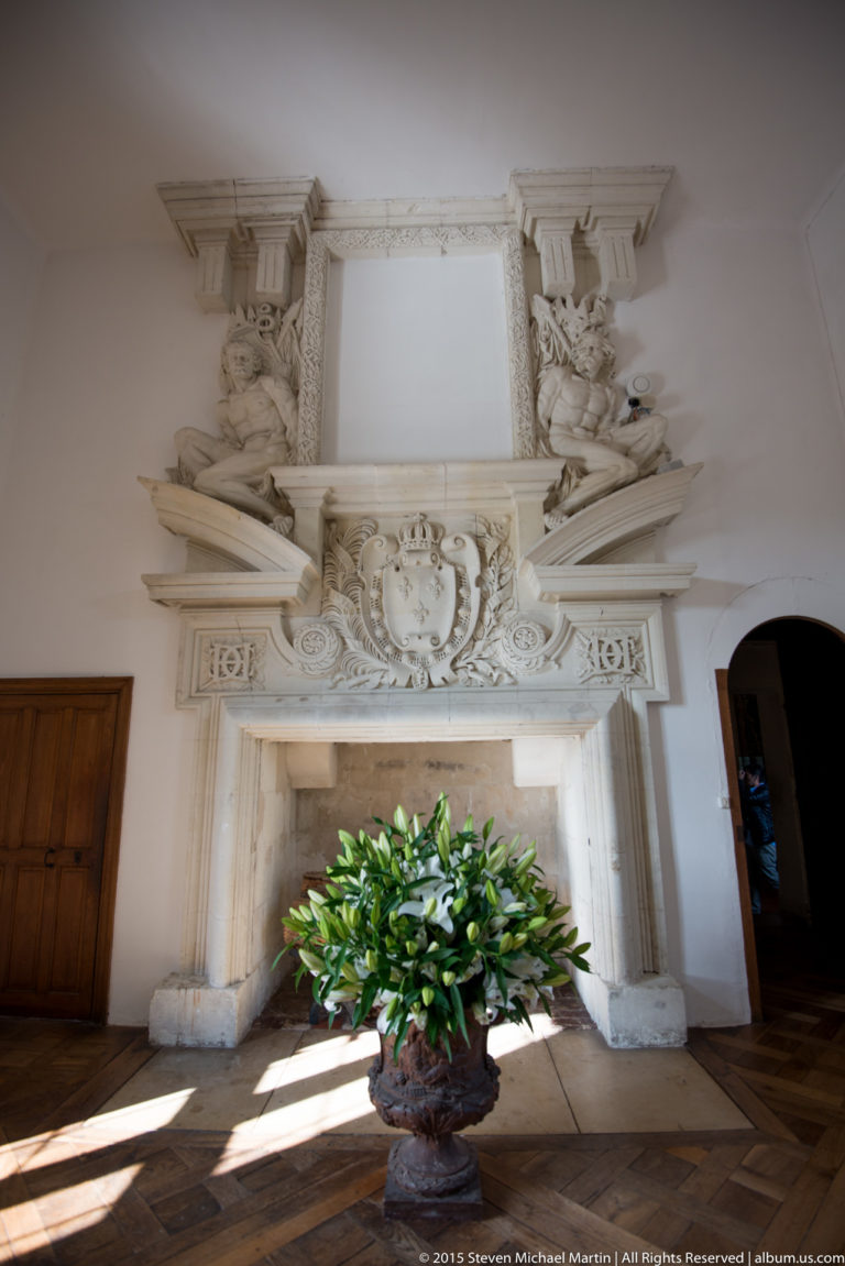 Large carved-stone fireplace and overmantel with sculpted figures, shield, scrollwork, and a potted plant at Château de Chenonceau.