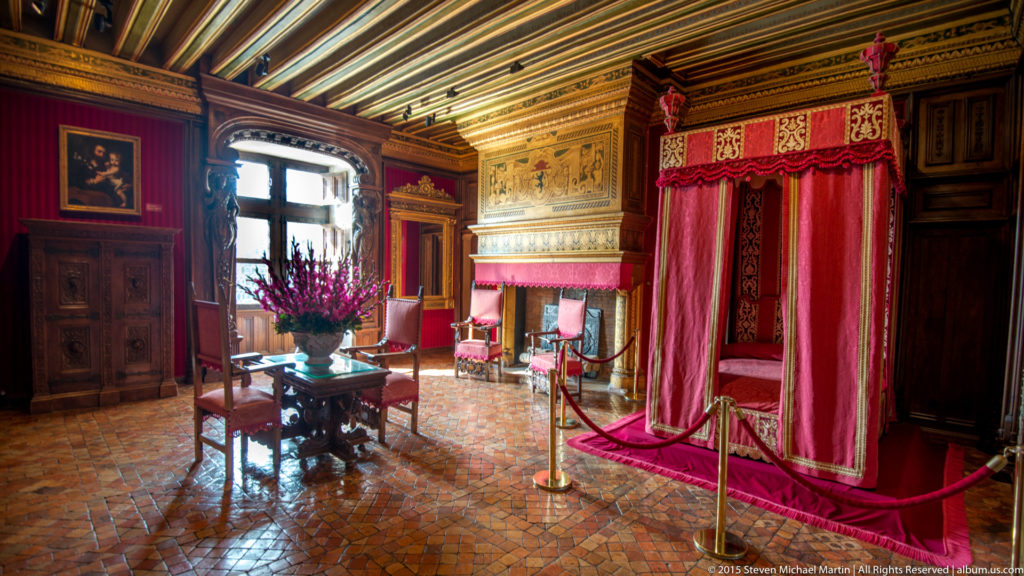 Wide view of a richly furnished bedchamber with red canopy bed, dark paneling, tiled floor, and floral table display at Château de Chenonceau.