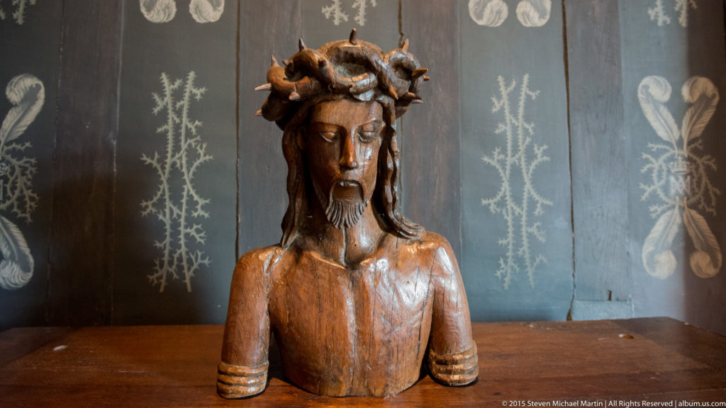 Carved wooden bust of Christ with downcast face, beard, and Crown of Thorns displayed against a dark wall.