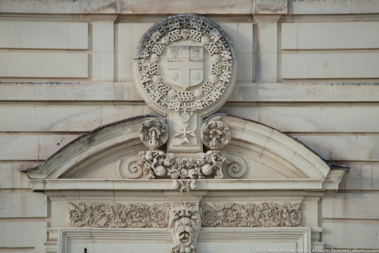 Stone heraldic cartouche above a doorway, with shield, carved chain, garlands, scrollwork, and a projecting human-face mascaron.