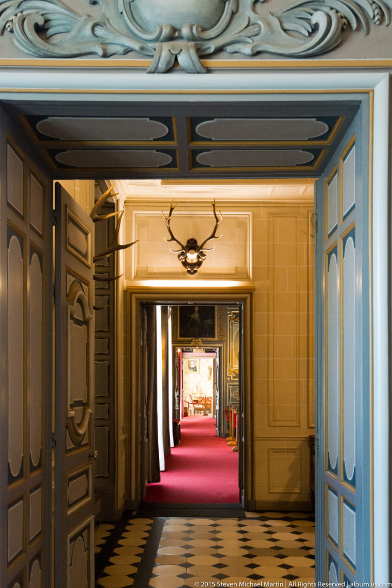 Aligned open doorways inside Château de Cheverny lead from a checkerboard floor through blue-gray rooms toward a red-carpeted interior.