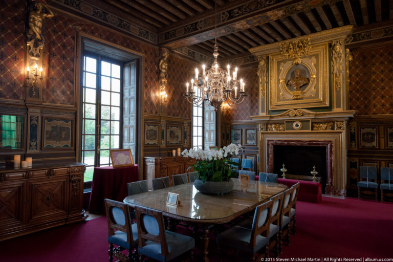 Formal château dining room with long table, carved furniture, patterned walls, painted panels, chandeliers, tall windows, and a fireplace.