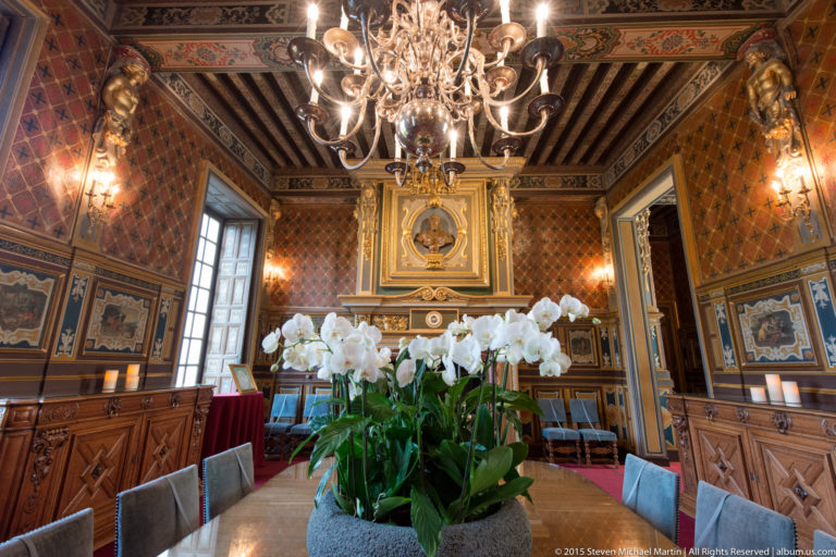 White orchids on a long dining table beneath a chandelier, with a fireplace, painted panels, and exposed beams in the background.