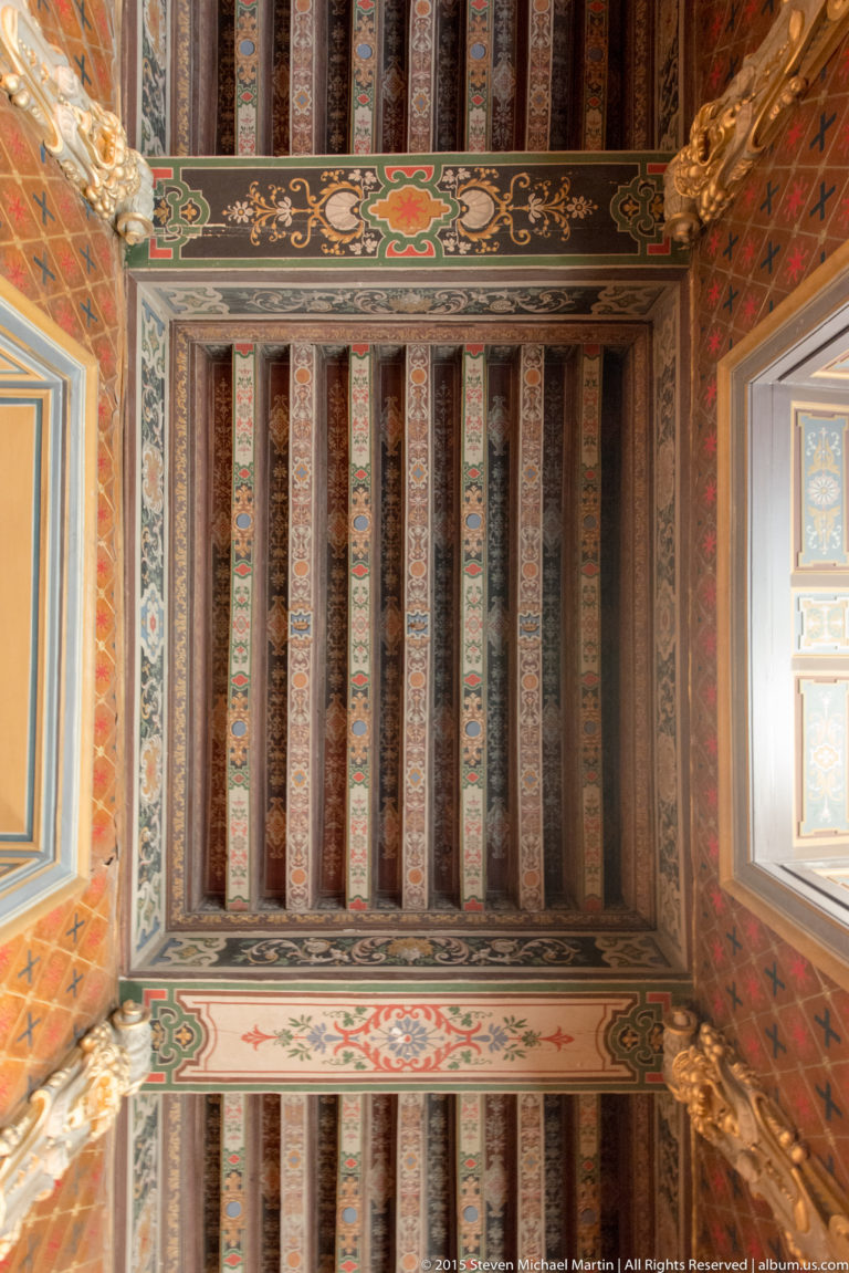 Upward view of a château ceiling with painted beams, colorful floral and geometric ornament, and bright window openings at both sides.