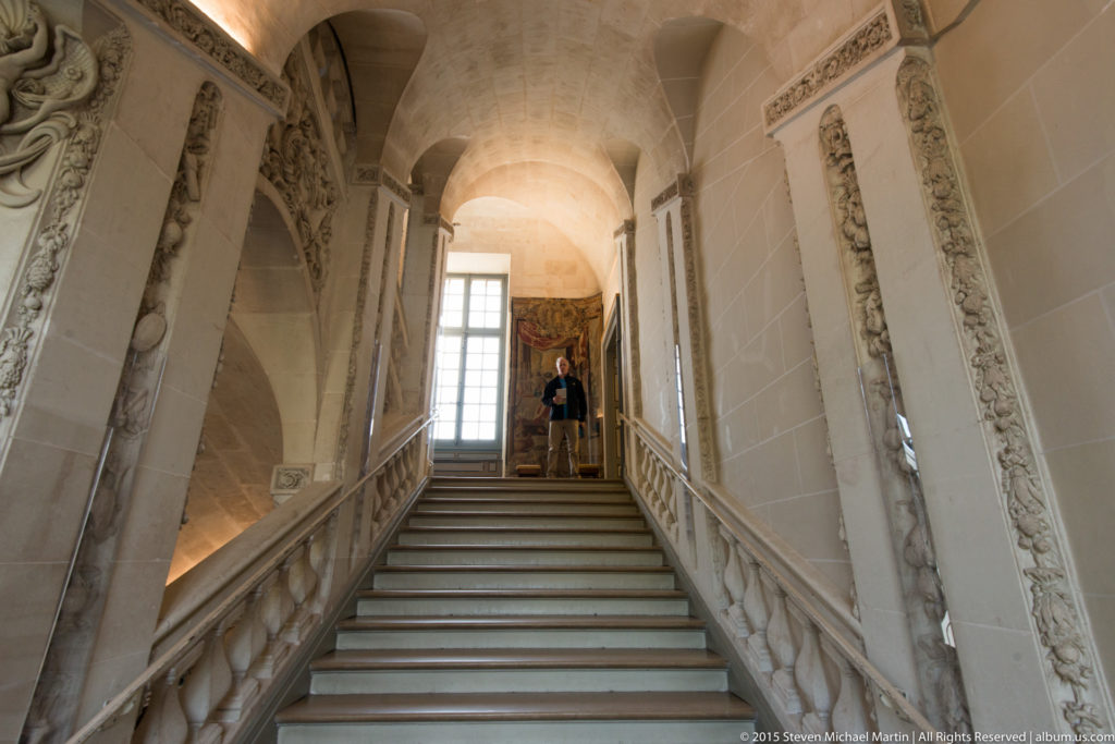 Pale stone staircase with carved balustrade, vaulted arches, wall reliefs, a tall landing window, and a visitor for scale.